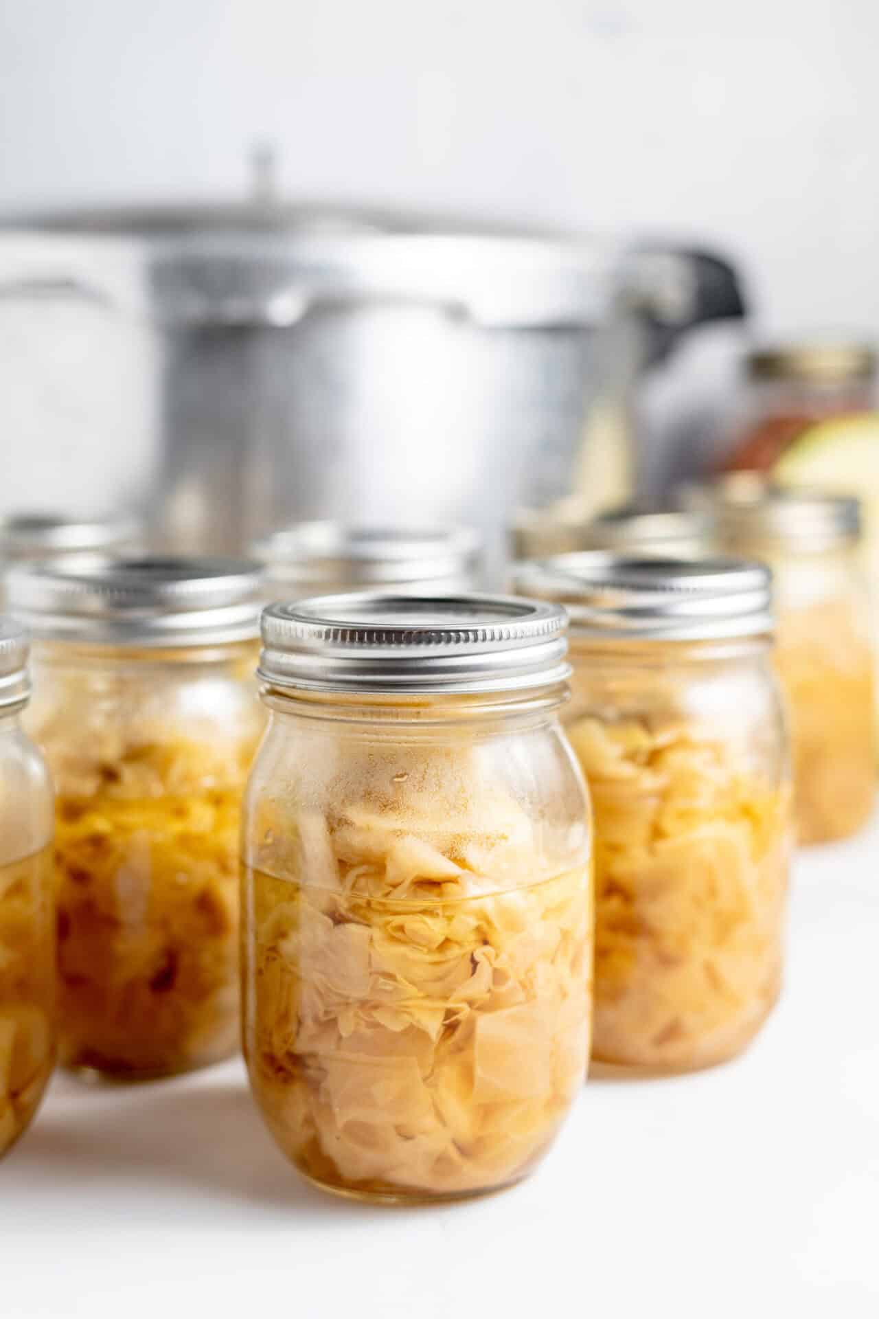 Image shows several rows of canned cabbage in glass jars lined up in front of a large stock pot. 