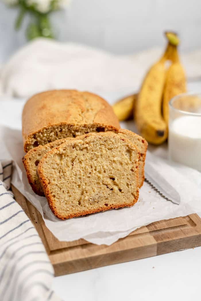Image shows sliced sourdough banana bread on a cutting board with a glass of milk and a small bunch of bananas in the background