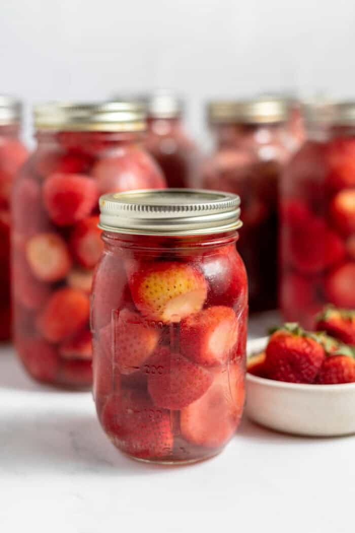 Image shows several jars of freshly canned strawberries with a bowl of strawberries next to it. 