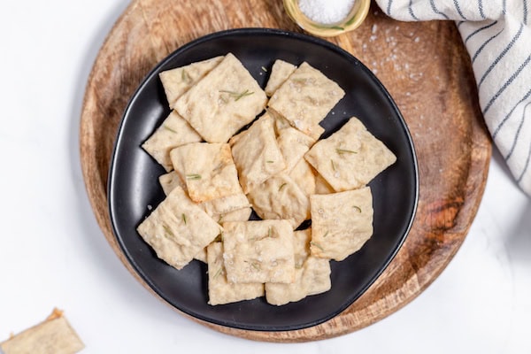 Image, taken from above, shows sourdough crackers with salt and rosemary on a black plate.