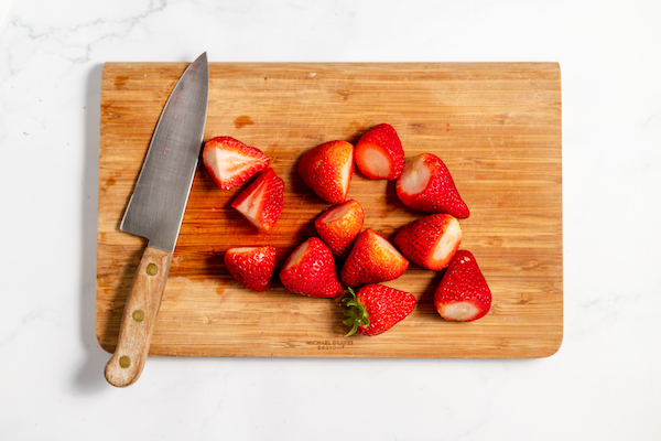 Image, taken from above shows sliced strawberries on a cutting board with a knife.