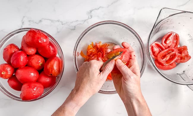 Image, taken from above shows a pair of hands using a knife to core and deseed Roma tomatoes