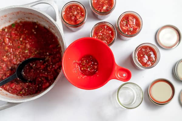Image, taken from above shows a spoon and funnel filling jars with fresh homemade salsa