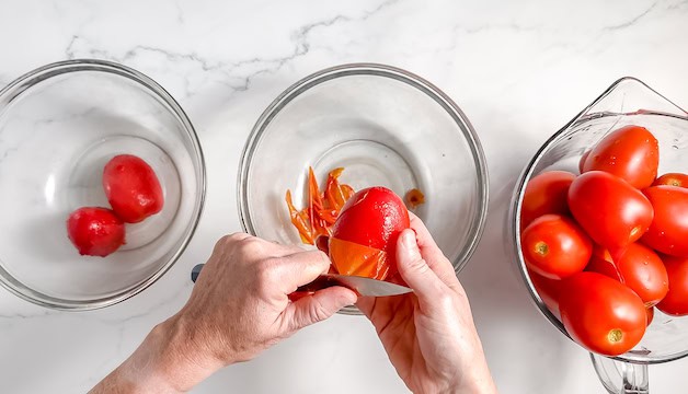 Image, taken from above shows hands using a knife to slip the peelings off of tomatoes into glass bowls