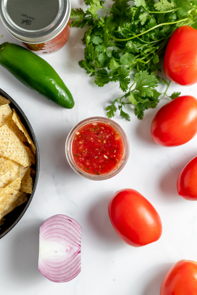 Image, taken from above shows a small jar of salsa surrounded by chips, and vegetables on table.