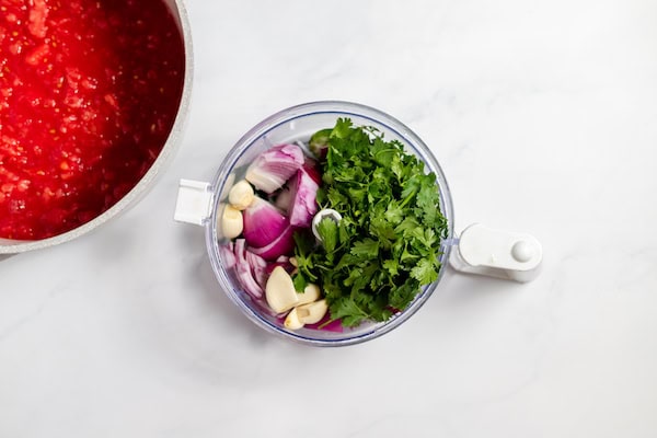Image, taken from above shows a bowl of tomatoes next to a food processor full of cilantro, garlic, onions, and jalapeño peppers.