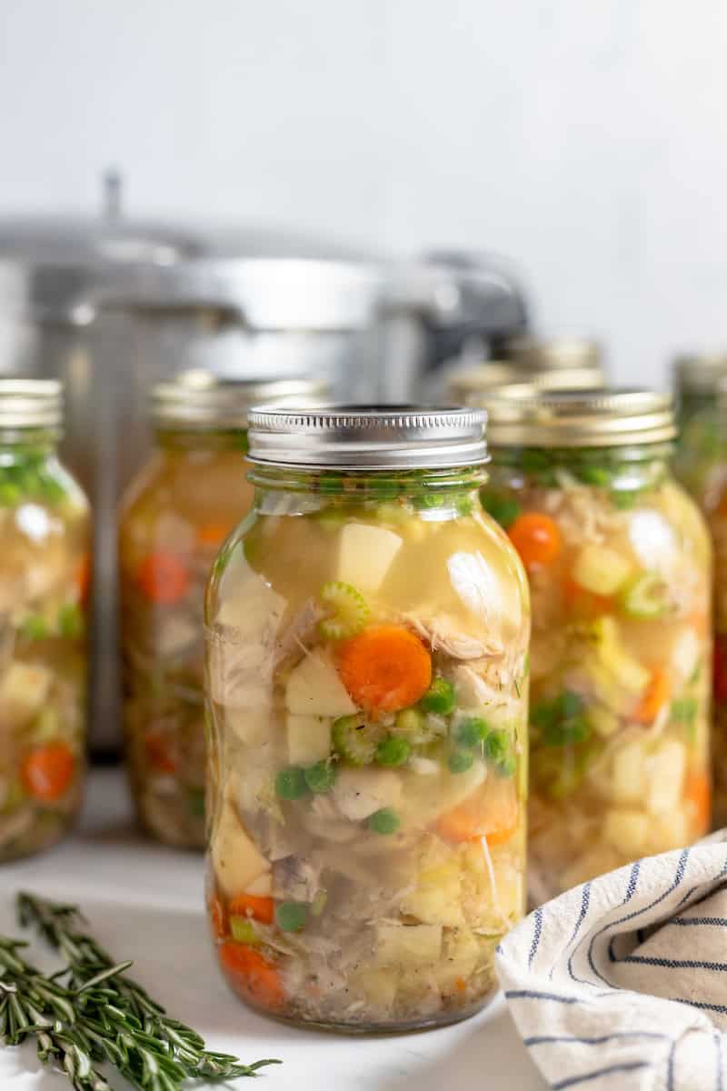 Image shows jars of chicken soup canned in glass jars, spread out on a counter top. 