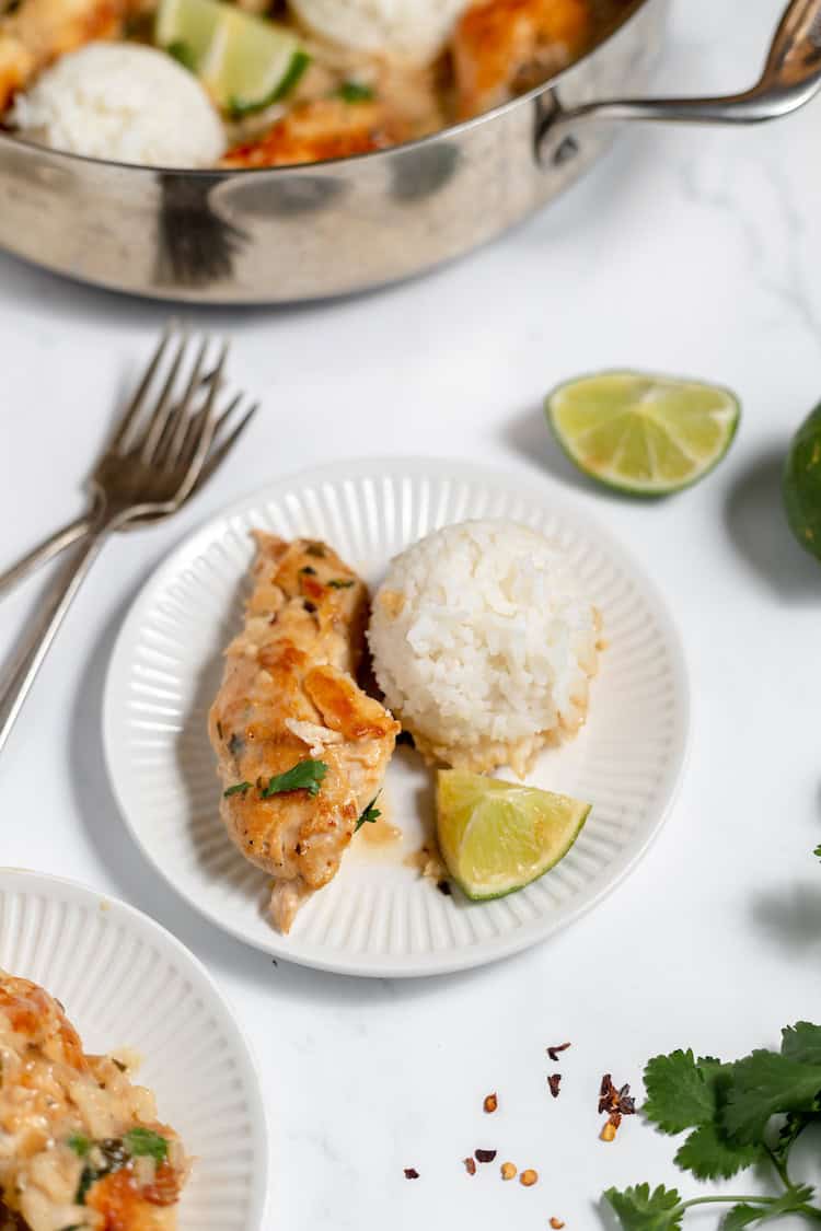 Image shows a small plate with a serving of rice, coconut chicken and a lime on it. Two forks sit nearby, along with the skillet full of the remainder of the meal. 