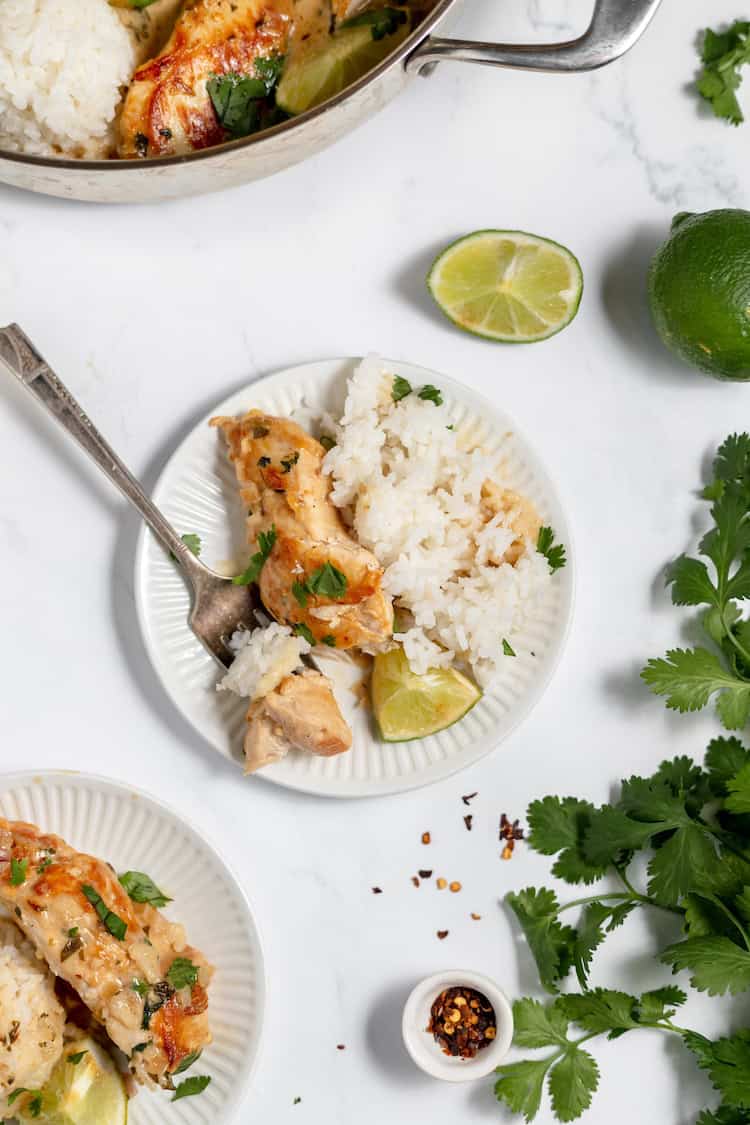 Image, taken from above shows a plate full of rice with lime and coconut chicken with a fork on the plate. The skillet sits nearby with more of the meal in it.