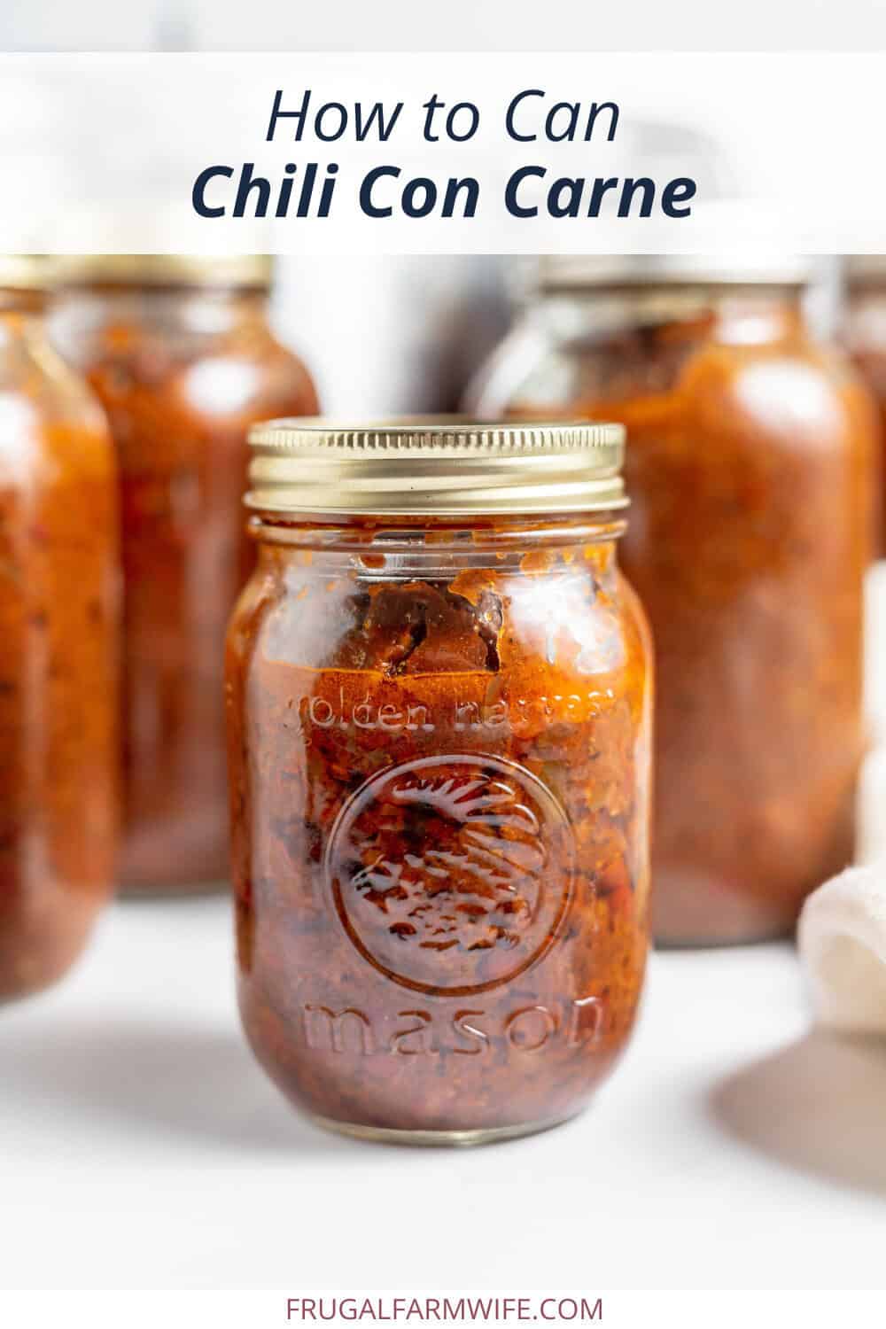Image shows a mason jar of chili con carne on a white counter top. Several more jars of chili sit behind it, with text above that reads "How to Can Chili Con Carne."