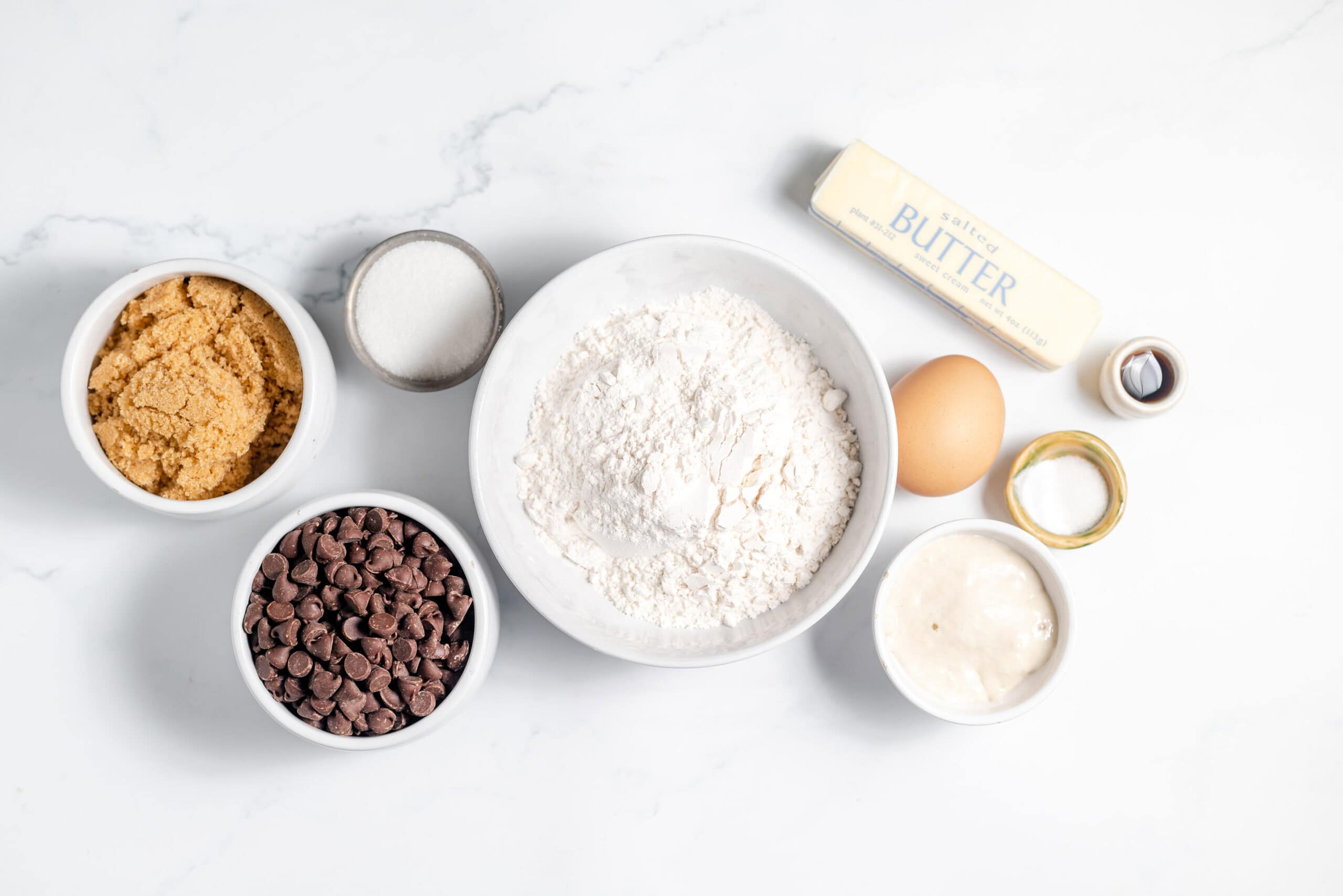 Image, taken from above shows several small bowls of the ingredients for making chocolate chip sourdough cookies laid out on a counter top. 