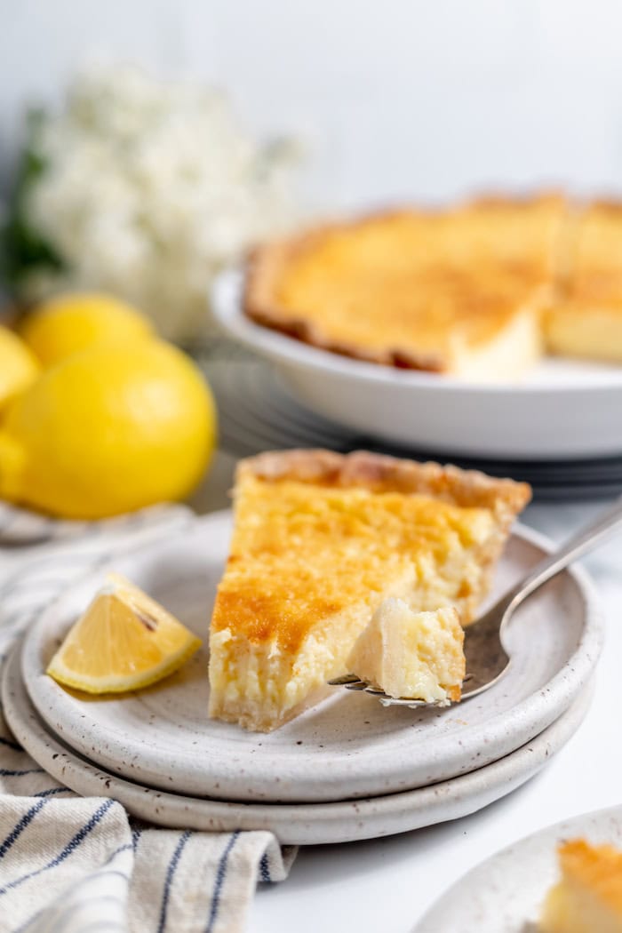 Image shows a plate with a piece of buttermilk pie with a bite taken from it. The rest of the pie sits in the background of the photo.