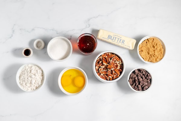 Ingredients, photographed from above, show several small bowls with the ingredients to make chocolate pecan pie bars, including eggs, butter, sugars and pecans.