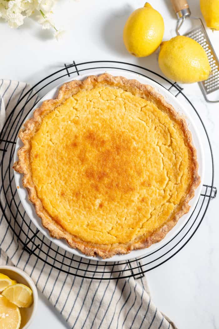 Image, taken from above, shows lemon pie filling in a sourdough crust on a wire rack. A few lemons are sitting nearby on the table. 