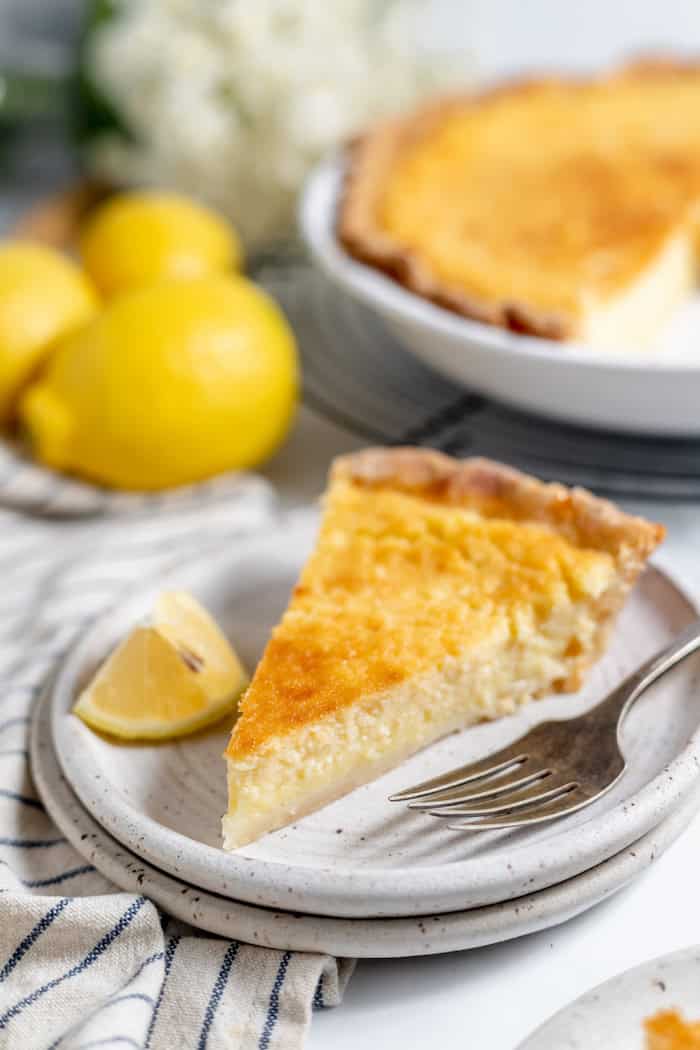 Image shows a plate with a slice of lemon buttermilk pie on it with a fork. Behind the plate is a dish with the rest of the pie. 