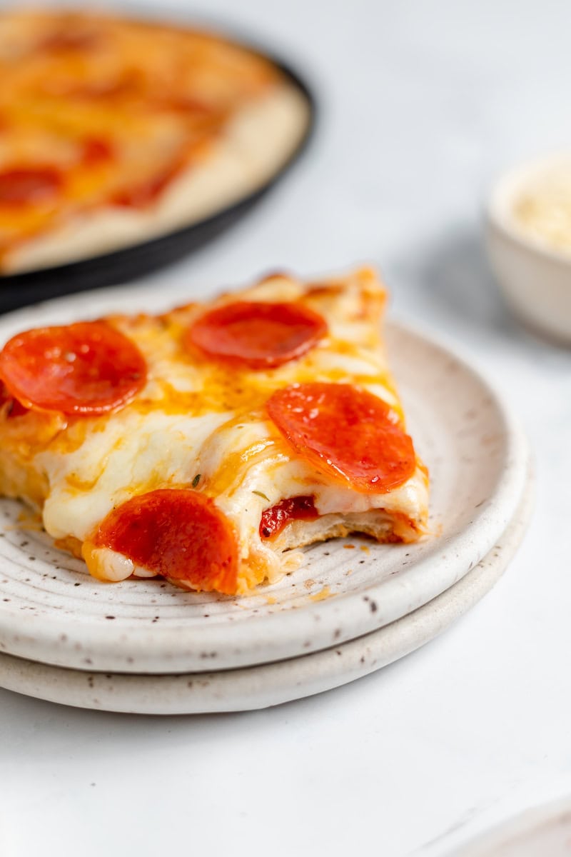 Image shows a slice of sourdough pizza with bite taken out of it on a plate. The rest of the pizza sits in the background of the photo. 