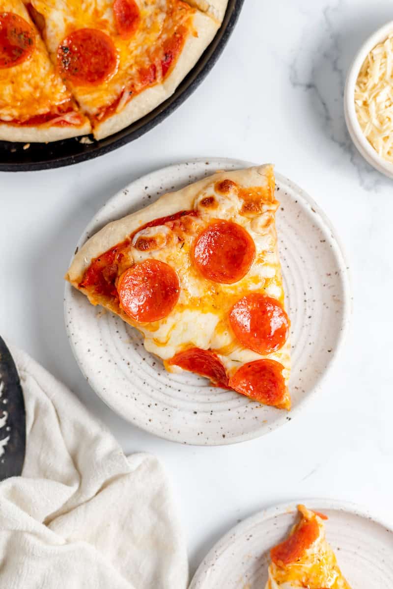 Image, taken from above, shows a slice of homemade sourdough pepperoni pizza on a plate. The rest of the pizza sits nearby. 