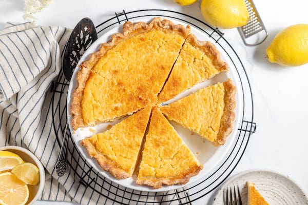 Image, taken from above shows a sliced buttermilk lemon pie on a wire rack. Several lemons are scattered nearby on the counter.