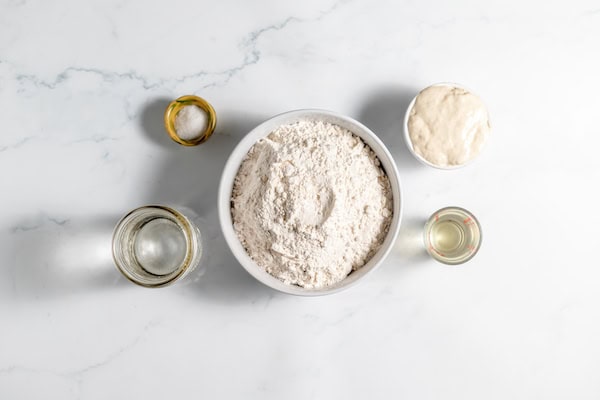 Image, taken from above, shows several bowls full of the ingredients for making sourdough pizza crust on a counter top