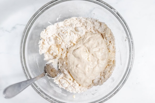 Image, taken from above shows sourdough starter added to flour in a glass bowl.