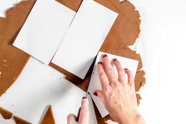 Image shows a pair of hands cutting pieces of gingerbread dough from the template shapes on rolled out dough.