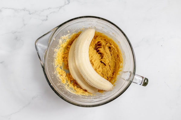 Image, taken from above, shows a mixing bowl with bananas added to butter and sugar mixture.