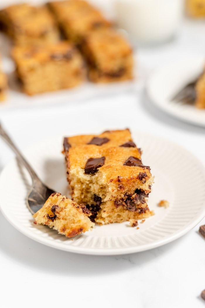 Image shows a chocolate chip banana bar on a plate with a fork. In the background a plate of more bars sits. 