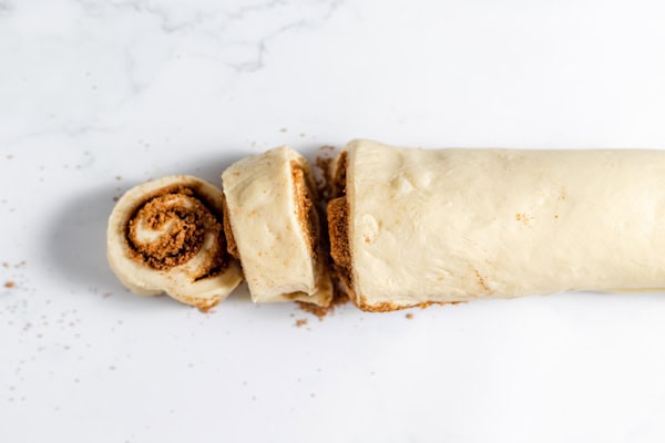 Image, taken from above, shows rolled up sourdough cinnamon rolls being cut into buns.