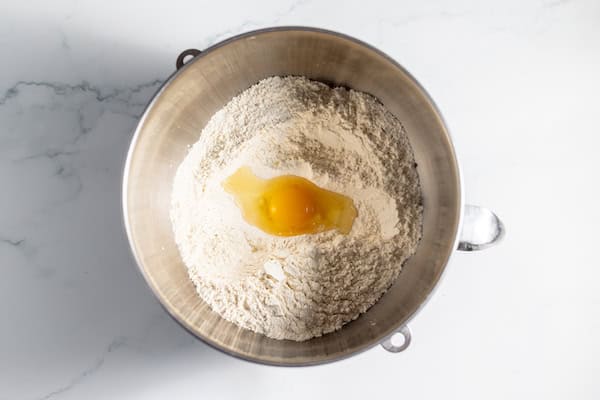 Image, taken from above, shows the dry ingredients for sourdough cinnamon rolls and egg in a glass mixing bowl.