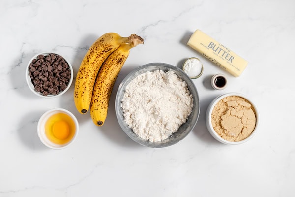 Image, taken from above, shows the ingredients to make chocolate chip banana bars laid out on a white counter. 