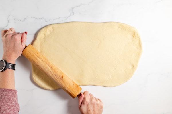 Image, taken from above, shows two hands rolling out sourdough roll dough on a white marble counter. 