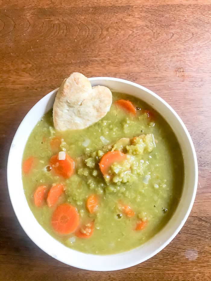 Image, taken from above, shows a bowl of split pea soup on a table with a small heart shaped cracker floating in it. 